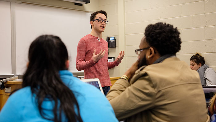 male student standing in front of classroom while peers listen to him speak