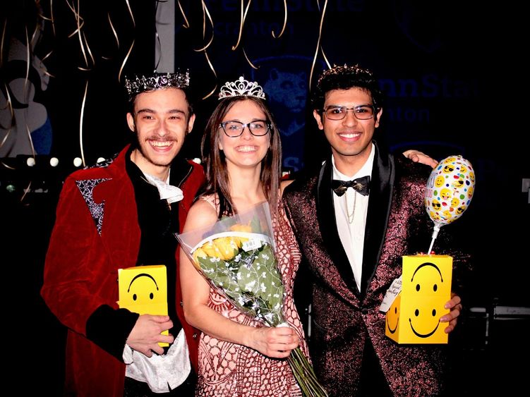 Three Student THON dancers wearing crowns and formal wear and holding yellow flowers pose for a photo after being announced at the THON Prom